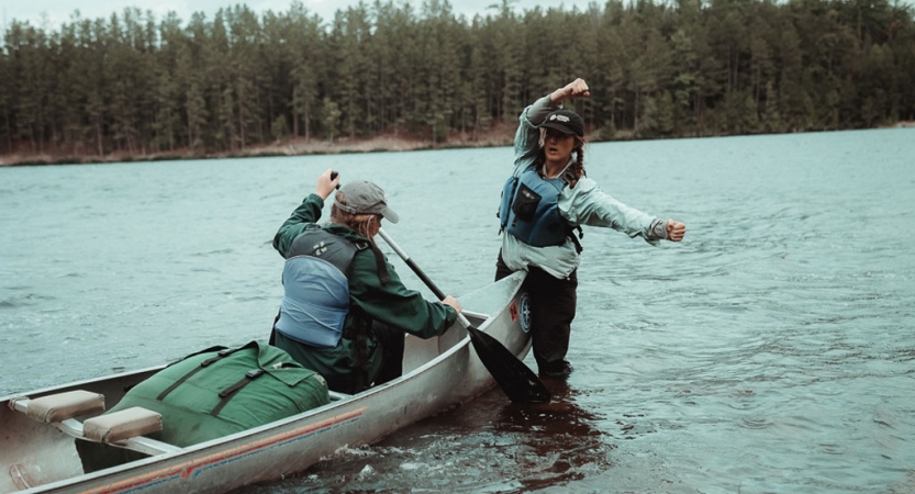 An instructor stands in the water, demonstrating how to paddle to a student in a canoe. They are both wearing life jackets. 